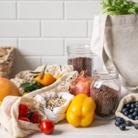 Fresh vegetables and fruits in eco cotton bags and cereals in glass jars on table in the kitchen. Zero waste shopping concept. Front view. Copy space.