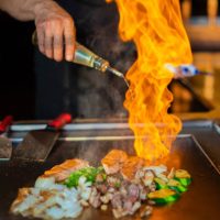 chef's hands with spatula over teppanyaki. cooking vegetables meat and seafood on hot hibachi grill table. Traditional Japanese Cuisine. Teppan show with flambe