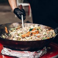 pasta with vegetables and seafood at street food festival. chef with gloves  cooking traditional italian macaroni dish. open kitchen outdoors, catering. summer picnic